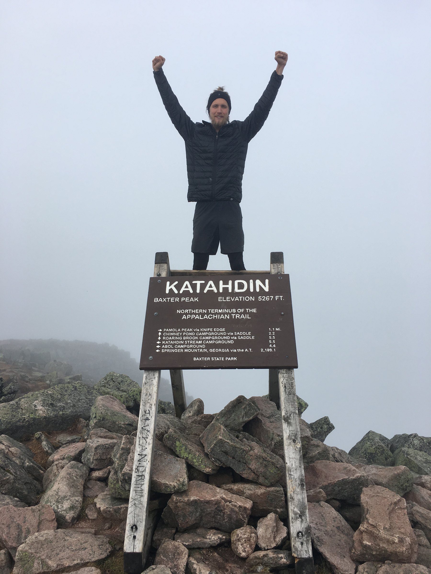 Jeremy standing with arms out beside a trail summit sign